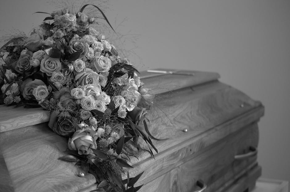 A detailed black-and-white photograph of a large, lush floral funeral tribute resting on a wooden coffin. The arrangement comprises a variety of flowers, predominantly roses in different sizes, with s