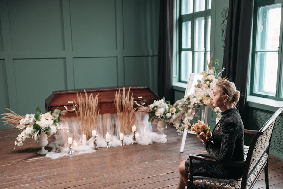 A woman sitting in a dimly lit room with green-paneled walls and large windows, holding a bouquet of peach and cream flowers. In front of her, there are two large floral arrangements in white urns, fe