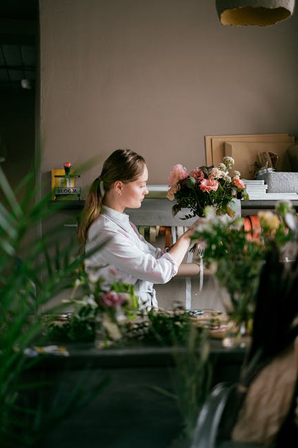 A young woman with light brown hair in a ponytail is arranging a floral bouquet inside a well-lit room. The bouquet features pink roses, white lilies, and greenery, wrapped in clear cellophane with a