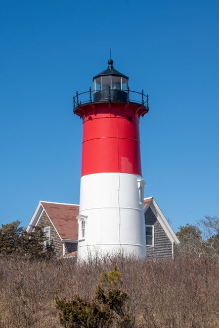 A tall, cylindrical lighthouse with a red upper section and a white lower section, topped with a black lantern room and a small balcony. The lighthouse is situated near a house with a brown tiled roof