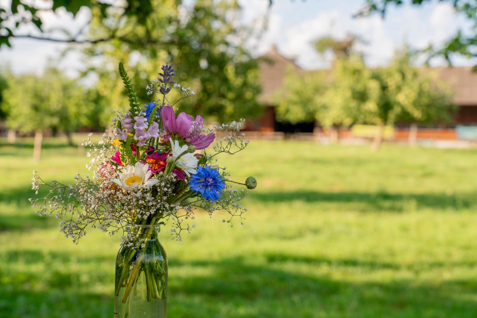 A clear glass vase holding a fresh floral bouquet featuring pink tulips, white daisies, blue cornflowers, and delicate baby's breath, arranged with green foliage. The bouquet is placed outdoors on a g
