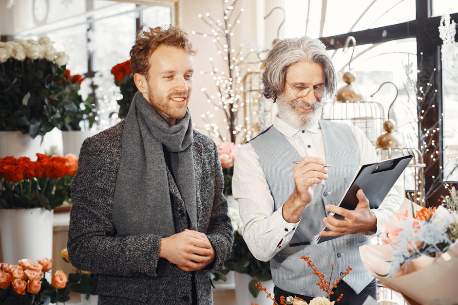 A young man with curly, reddish-brown hair and a beard stands beside an older male florist with gray hair and a beard, both inside a well-lit flower shop filled with various fresh flowers, including r