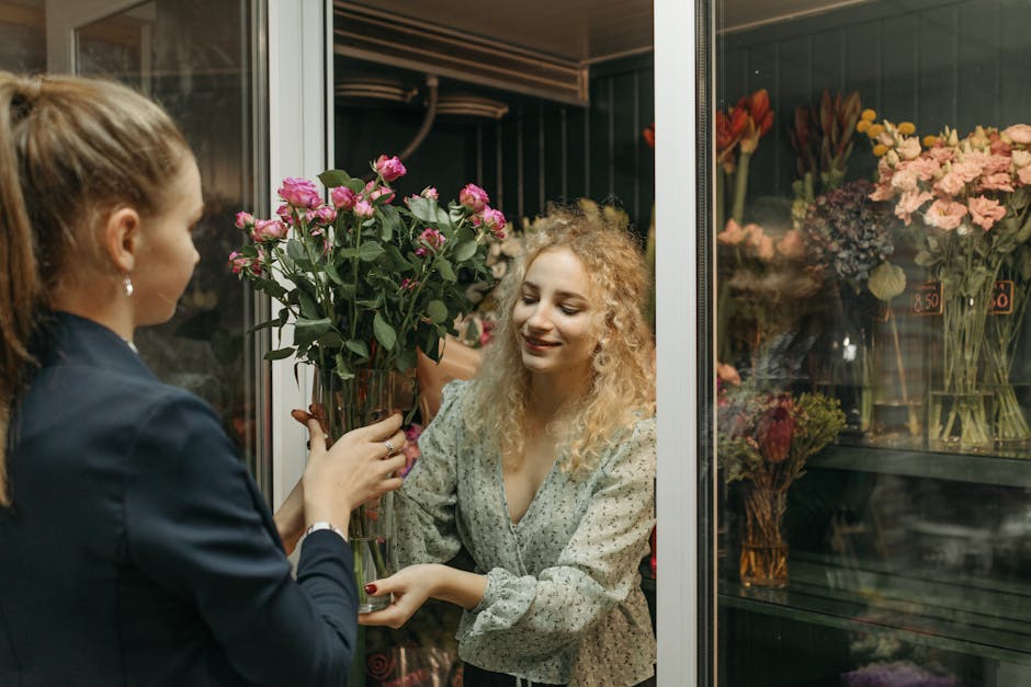 A florist at Floristeastham.co.uk is handing a tall, clear glass vase filled with freshly cut pink roses to a smiling female customer through the glass door of a floral shop. The roses have lush, vibr