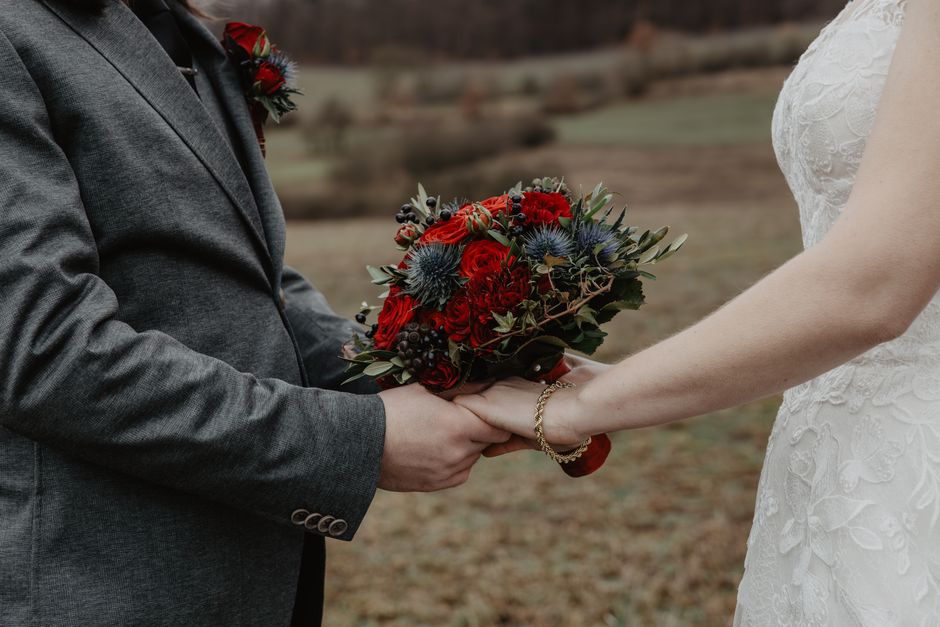 Close-up of a couple during a wedding, with the groom in a grey suit holding a vibrant bouquet of red roses, blue thistles, and dark berries, part of a floral arrangement gifted as a wedding bouquet.