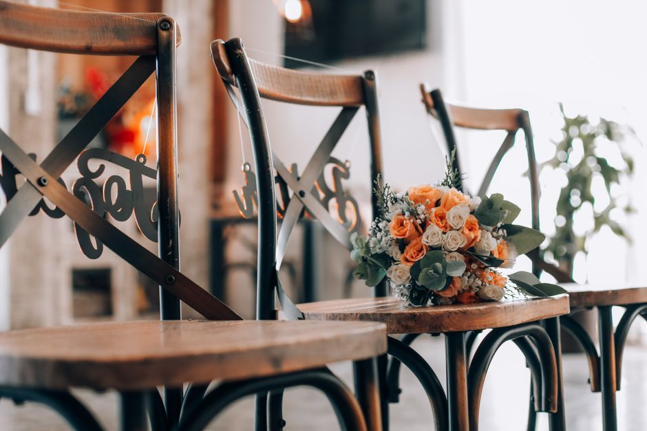 A bouquet of fresh floral arrangement resting on a wooden chair, featuring white and peach roses, complemented by sprigs of white baby's breath and green eucalyptus leaves. The bouquet is wrapped in c