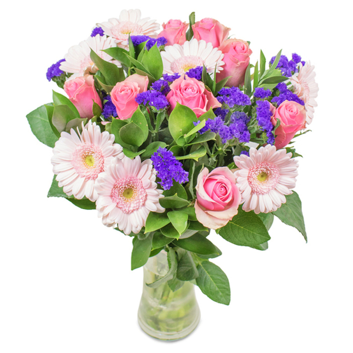 An elegant and vibrant floral arrangement sits gracefully in a tall, clear glass vase-a distinctive creation from Florist East Ham, reflecting the diversity and freshness of the local neighbourhood. The centrepiece features an inviting blend of approximately seven to nine rich pink roses, with several still unfurling in perfect spiral-symbolic of classic romance and new beginnings. Scattered among them are five to seven pale pink gerbera daisies, their large circular faces and slender petals offering both texture and lightness, each centre glinting softly as if capturing the morning sun streaming through a window on High Street North. Interwoven throughout the bouquet are clusters of vivid purple statice, adding a lively contrast against the soft pastels and echoing the vibrancy found at the heart of East Ham's vibrant markets. The deep green foliage below provides a lush, abundant base, enhancing both the sumptuousness of the bouquet and the sense of fresh possibility. Visible stems and water evoke a sense of authentic freshness, perfect for local delivery to mark a birthday, anniversary, or to elevate everyday life with a touch of East Ham's distinctive charm. This arrangement offers both visual delight and emotional resonance, making it a thoughtful choice for home, office, or heartfelt gift.